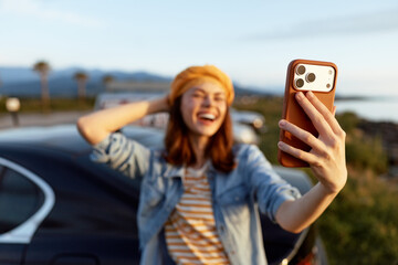Naklejka premium A cheerful woman takes a selfie outdoors beside a car, enjoying sunny weather. A lifestyle portrait capturing spontaneous joy, confidence, and carefree moments.