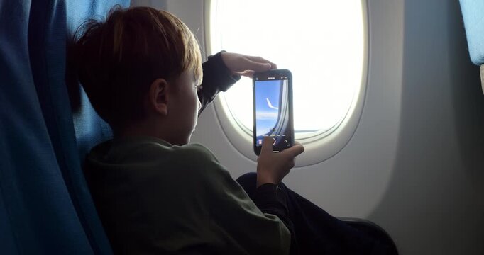 Young passenger capturing video through airliner window using smartphone, focused on airplane winglet. Slow motion shot, boy documenting travel experience with curiosity and concentration