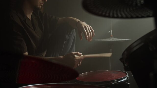 Young man wearing wireless earbuds sitting by drum kit in dark studio, holding drumsticks and practicing rhythm. Ideal for music education and audio equipment commercials