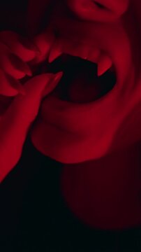 Vertical video. Close Up View of Young Woman Placing Artificial Vampire Fangs Into Mouth Under Red Light, Halloween Costume Preparation, Horror Style, Festive Transformation