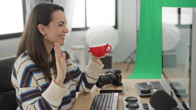 Woman holding a red mug with hands visible, smiling at monitor with camera and keyboard on desk in studio; cheerful productivity.