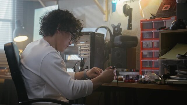 Asian young adult man soldering circuit board at workshop desk with computer tower in background. Clip is ideal for tech repair services, engineering education, and hardware development promos