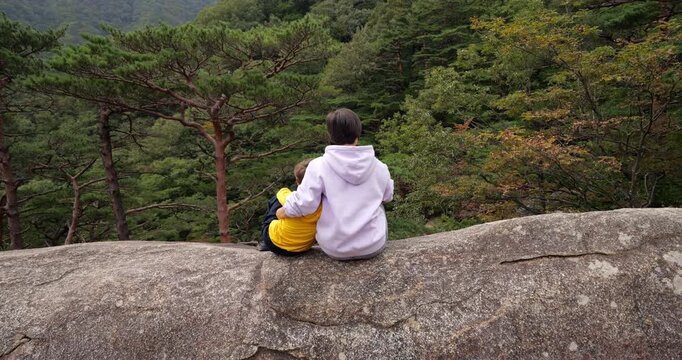 Loving mother and son share quiet embrace on rocky cliff, gazing at expansive forested mountain landscape during peaceful autumn journey. Slow motion shot, view from behind