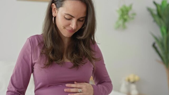 Pregnant woman wearing mauve top gently caresses her belly while seated on a bed in a bedroom, smiling and looking down; maternal serenity.