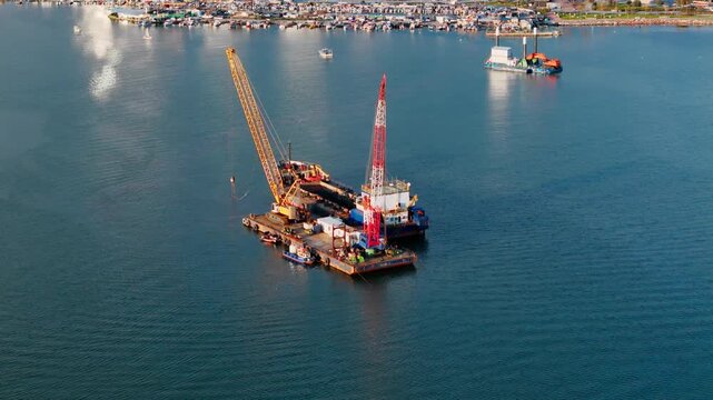 Floating crane barge beside dredging ship during coastal water maintenance work. Aerial view of industrial marine operation