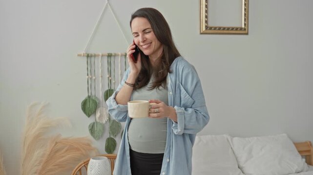 Pregnant woman talking on smartphone while holding a coffee mug and exposed belly in a bedroom with macrame wall hanging and pampas grass; anticipation motherhood.