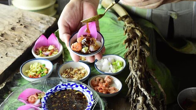 Person pouring sweet savory sauce over Miang Kham traditional Thai appetizer with pink lotus petals and herbal ingredients, presenting authentic Southeast Asian cuisine lifestyle.