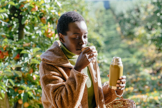 Thoughtful black woman drinking hot tea from thermos in sunny apple orchard