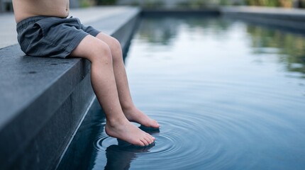 Fototapeta premium Little boy sitting on the edge of a swimming pool with feet touching the water