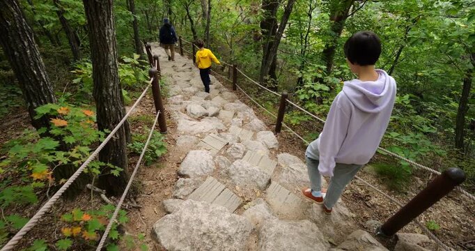 Tourist family descending steep stony path after visiting Ulsanbawi Rocks, walking spread out with father leading, energetic son following, and mother behind using tree trunks for support, follow shot