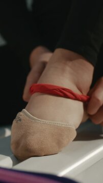 Close Up of Rhythmic Gymnast Foot in Specialized Half Socks Inside Changing Room. Athlete Ties Red Elastic Resistance Band Around Arch and Ankle, Likely For Injury Prevention, Support. Vertical Shot.