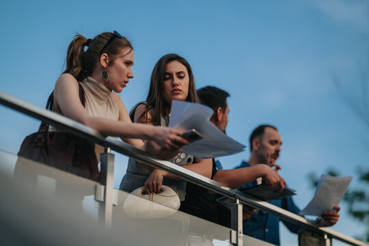 Business colleagues stand on a balcony reviewing papers and a smartphone in a focused discussion. A small group rechecks documents and discusses plans during an outdoor work meeting.