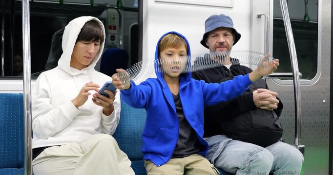 Young boy sits between his parents on subway train, swinging and stretching shiny metal spring toy, while his father watches attentively and his mother remains focused on her smartphone.