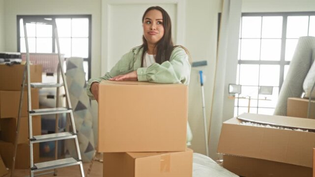 Woman leaning on a large cardboard box, smiling and resting her hand on cheek amid packed moving boxes, ladder, rolled mattress and mop in a sunlit building room; joy new beginnings.