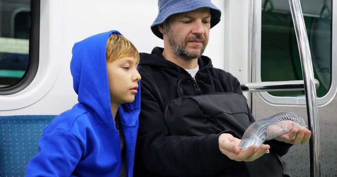 Father and son sitting side by side on subway train, as man playfully stretches and swings metal metal spring toy between his hands. boy, once curious, now watches in quiet fascination.