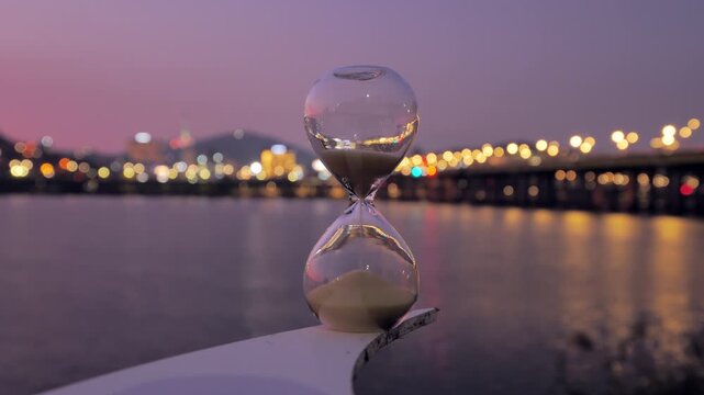 Sand cascading through hourglass bulbs, symbolizing time's progression, with blurred urban lights of Seoul glimmering on the river surface during twilight's ethereal moments.