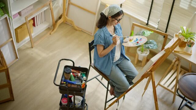 High angle view of Young Asian woman artist painting on canvas easel. Talented female painter working in bright home art studio or cozy living room. hobby, relaxation, and artistic lifestyle concept.