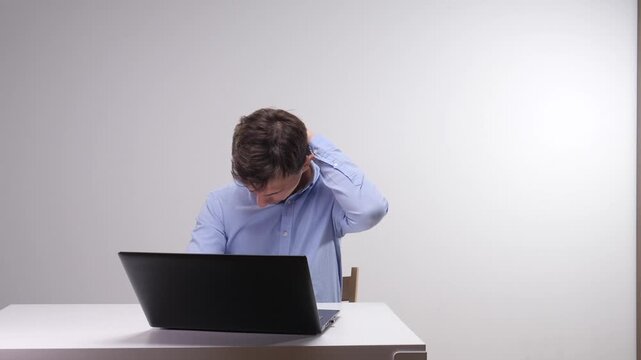 Young man working at a laptop feeling pain in his neck, rubbing it with a hand due to prolonged sedentary work and bad ergonomics causing strain and discomfort