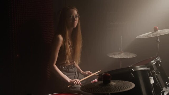 Portrait of Caucasian young womanr with glasses sitting at drum kit in music studio, holding drumsticks and smiling. Footage for music school promotions or youth hobby advertisements