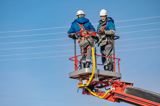 Two electricians in safety gear are working on power lines from a lift. They are focused on their task against a clear blue sky.