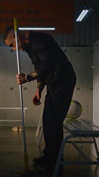 Cleaner in Takes Break From Mopping Locker Room. Man Sits on Bench, Picks Up Soccer Ball Left Behind by Team, and Holds it With Both Hands, Contemplates Youth With Sense of Nostalgia. Vertical Shot.
