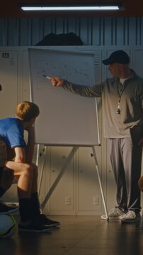 Middle Aged Soccer Coach in Black Cap Stands by Whiteboard, Drawing Game Tactics, Explaining Strategy to Teenage Players. Youth Football Team Sits on Benches, Focused on Presentation. Vertical Shot.