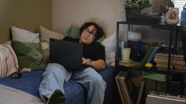 Young adult Asian man lying on bed and working on laptop, looking tired. Ideal for commercial use in remote work fatigue, online education, or freelance burnout campaigns, zoom out