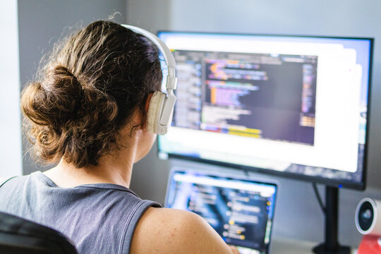 Man wearing wireless headphones works at a desk with laptop and external monitor in a home office. Concept of remote work, software development, productivity and focused concentration.