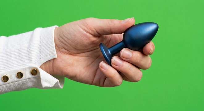 A person's hand wearing a white long-sleeve shirt holds a metallic blue butt plug against a solid bright green background.