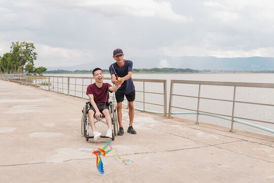 Happy young disabled man in wheelchair laughing while flying a colorful parrot kite with male caregiver on a lakeside walkway, inclusive outdoor lifestyle, accessibility and support concept.