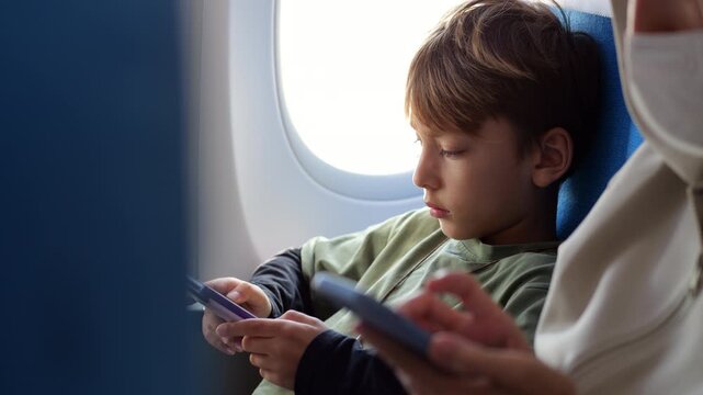 Passenger boy deeply focused on his electronic book while flying, seated by window next to his mother, who is using smartphone, seen blurred at foreground. Modern family travel moment