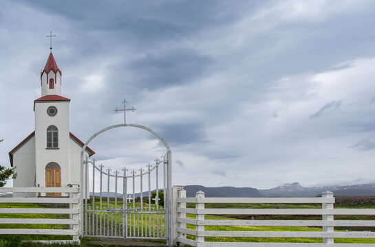 White church and metal cemetery gate with crosses standing against a dramatic cloudy sky, with green grass and distant mountains defining the remote landscape