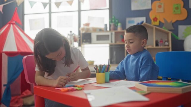 Girl and boy sit at a red table drawing, girl holds pencil at chin in a thinking pose while boy reaches into a cup of colored pencils in a classroom; teamwork learning focus.