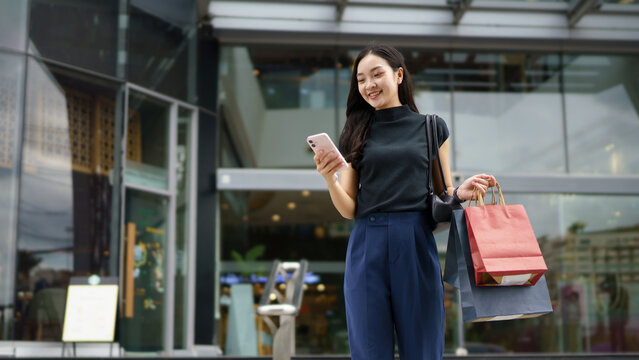 Smiling young woman shopper customer standing on city street holding shopping bags and smartphone. Sale e commerce app. Consumerism, black Friday. Online shopping.