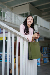 Fototapeta premium Happy Young Asian Woman Holding Shopping Bag and Coffee Cup Leaning on White Railing at Shopping Mall