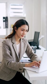 Young asian professional business woman working on laptop, typing using computer looking at screen searching information at office.