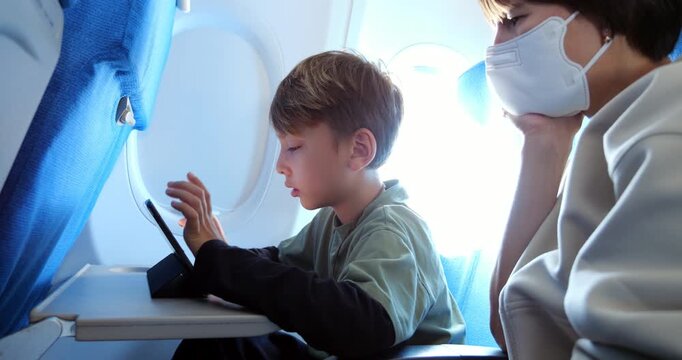Child passenger enjoying educational game on tablet during flight, solving tasks and speaking while seated next to his mother, who watches supportively and occasionally offers comments.