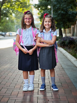 Two Happy Schoolgirls in Uniforms with Backpacks Ready for Back to School. Smiling Sisters Standing Together Outdoors on First Day of Primary School.