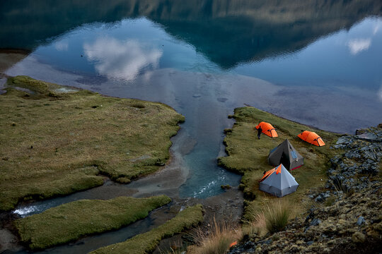 a small cluster of orange and grey expedition tents occupies a narrow tongue of vivid green moss beside a glacial stream where it spills from Carhuacocha lagoon into the valley below. 