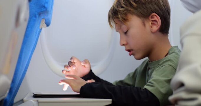 Young passenger boy seated at window seat with shade closed, growing tired while using educational app on his tablet during flight. Despite rubbing his eyes and showing signs of fatigue