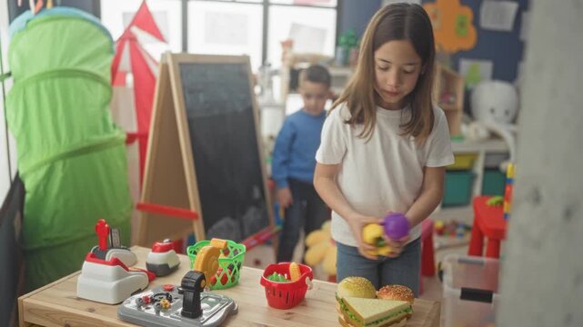 Girl and boy sorting toy food, baskets and play kitchen items on a wooden table in a preschool building, hands arranging plastic fruit and sandwich; calm concentration.