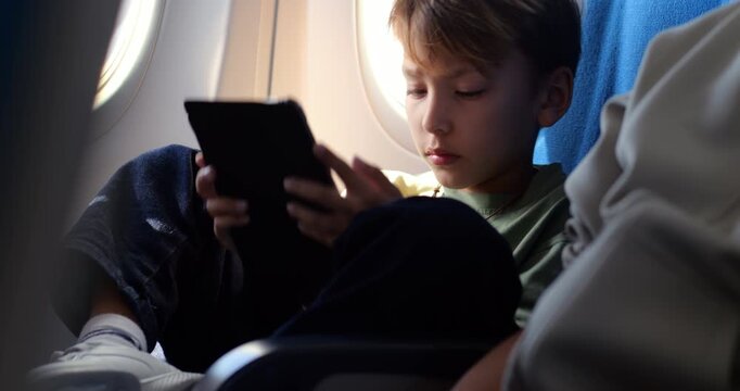 Young passenger reading digital book on his tablet while sitting near airplane window with legs crossed up on seat during flight. Boy enjoys quiet entertainment and peaceful journey
