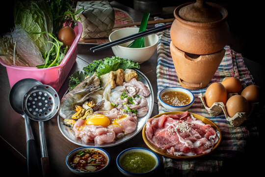 Traditional Thai clay pot hot pot known as Jim Jum, featuring raw sliced pork, fresh seafood, egg, vegetables, and spicy dipping sauces on a wooden table, perfect for an Asian food concept. 