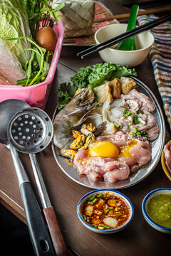 Traditional Thai clay pot hot pot known as Jim Jum, featuring raw sliced pork, fresh seafood, egg, vegetables, and spicy dipping sauces on a wooden table, perfect for an Asian food concept. 
