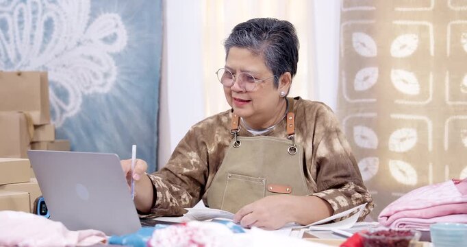 Senior asian woman businesswoman writes invoice notes beside laptop, checks online order list, compares shibori fabric bundle, stacks parcel box, prepares packing sequence for shipping pickup.