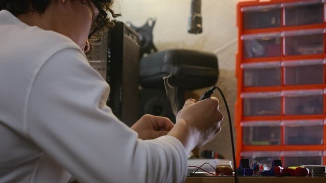 Asian young adult man soldering electronic components on circuit board with smoke rising. Video is ideal for engineering education, DIY hardware repair tutorials, or tech startup promotions