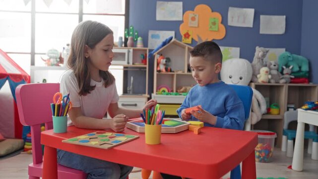 Girl and boy play with a wooden shape puzzle and colored pencils at a red table inside a building; concentration learning cooperation.