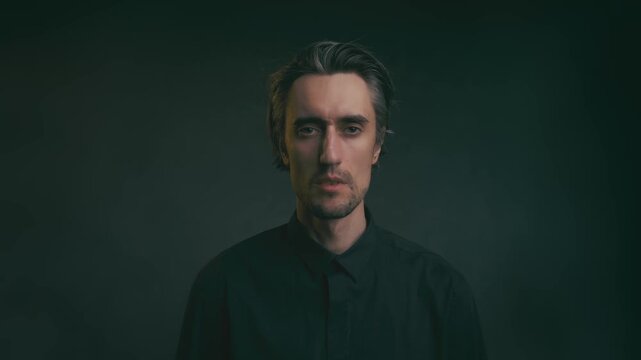 Moody studio portrait of adult man with medium-length dark hair and goatee in black collared shirt, looking at camera against dark gradient background