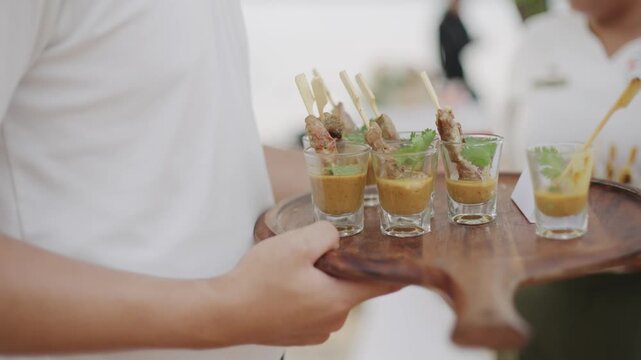 A server carrying a wooden tray with delicious meat skewers in small glass cups, perfect for catering, cocktail parties, and luxury hospitality events.