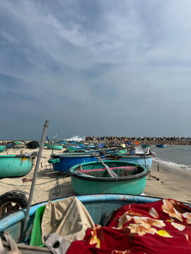 Colorful coracle basket boats sit on the shore under a blue cloudy sky. An ocean breakwater stands in the background as waves gently hit the sandy coastline.